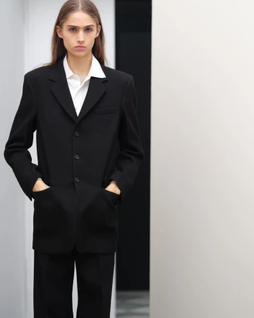 A young adult woman wearing a black suit with a white shirt poses indoors against a plain wall in a portrait-style fashion shot.