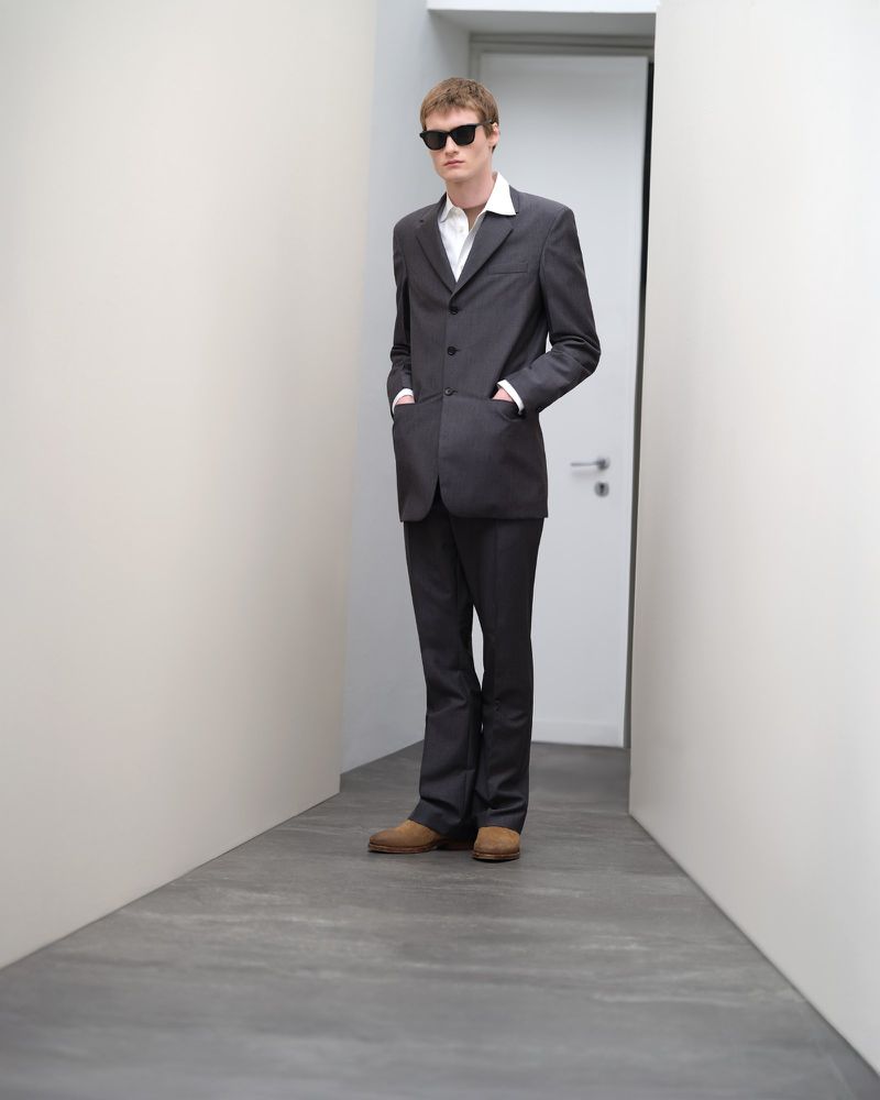 An adult male wearing a dark suit and sunglasses stands in a brightly lit hallway with neutral walls and flooring in a full shot.