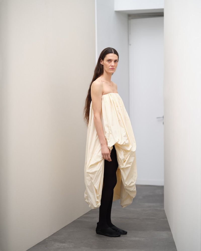 A solitary woman poses indoors wearing a voluminous, cream-colored strapless dress with black leggings against a neutral hallway.