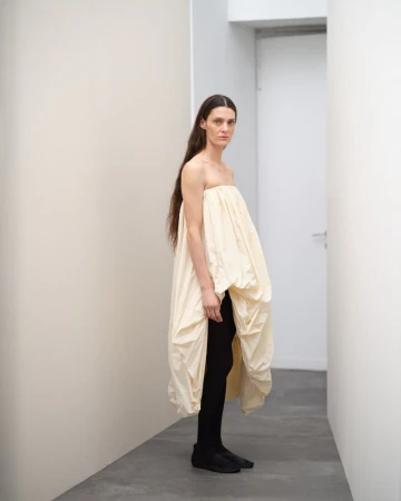 A solitary woman poses indoors wearing a voluminous, cream-colored strapless dress with black leggings against a neutral hallway.