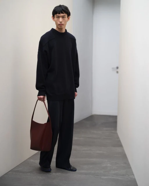 A slender Asian man, dressed in a dark top and slacks, poses indoors holding a maroon hobo bag against a neutral modern background.