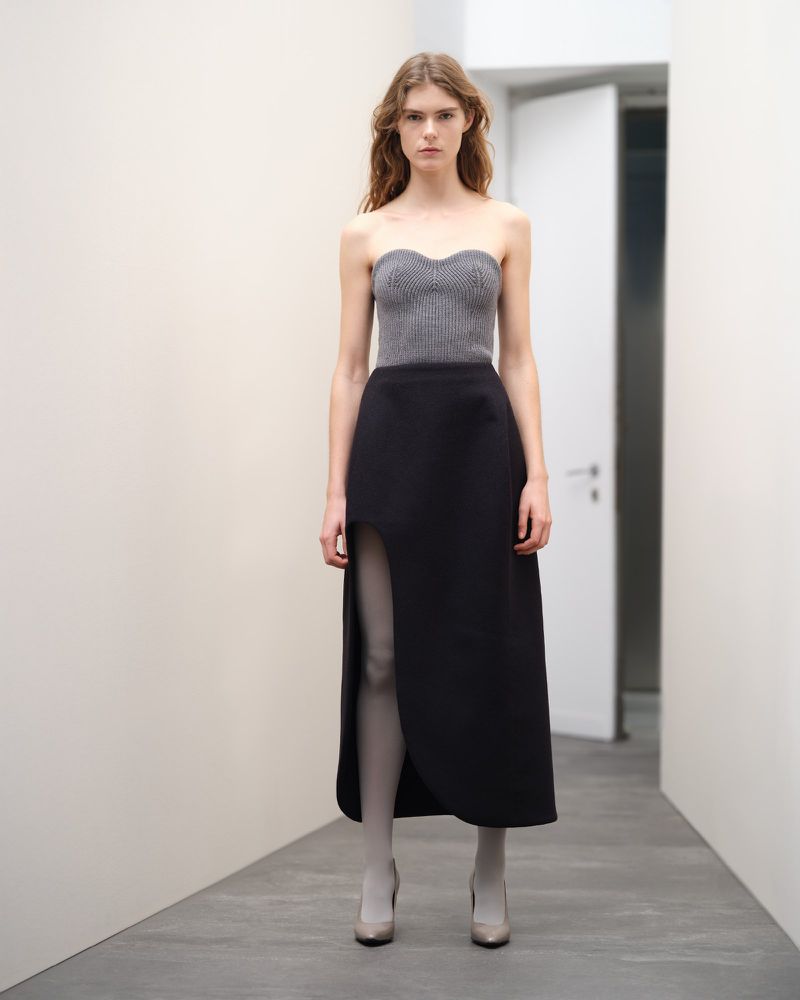 Full-length fashion portrait of a young adult woman in a gray strapless top and black slit skirt, posing in a minimalist white interior setting.