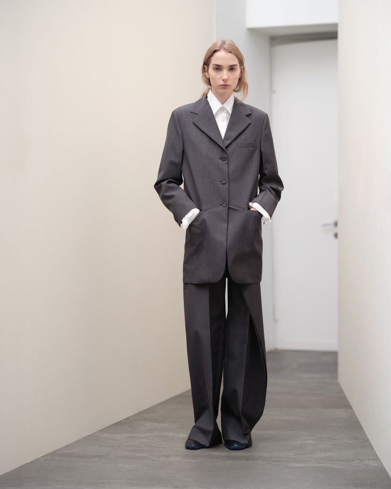 Blonde woman in gray, oversized blazer and pants stands in a hallway with hands in pockets, looking directly at the camera in a fashion shot.