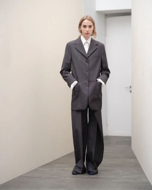 Blonde woman in gray, oversized blazer and pants stands in a hallway with hands in pockets, looking directly at the camera in a fashion shot.