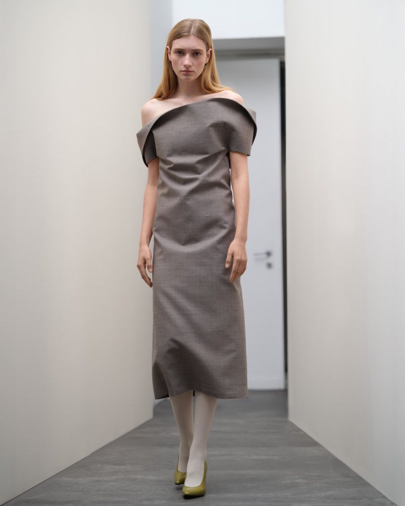 A blonde woman is posing in a studio in a gray, off-shoulder dress with a midi skirt, white tights, and green heels, looking forward.