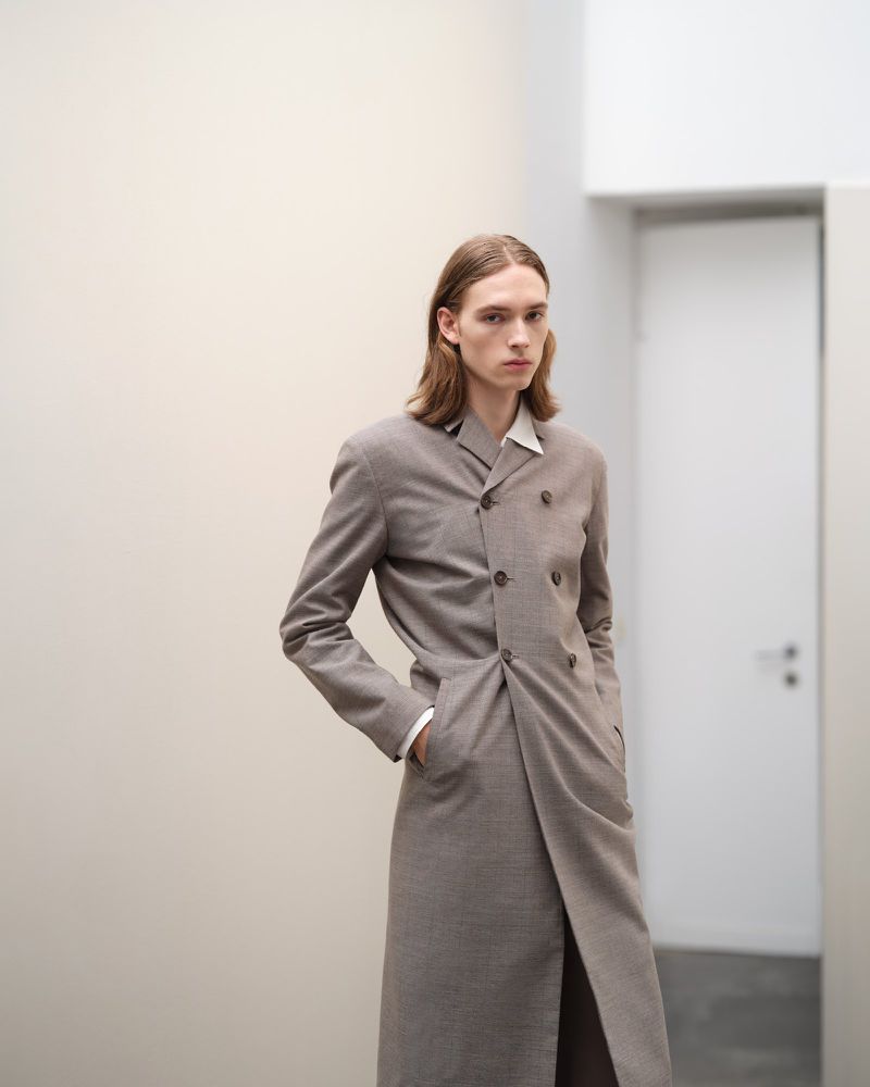 Model wearing a long taupe double-breasted coat standing in a minimalist interior space for Jil Sander.