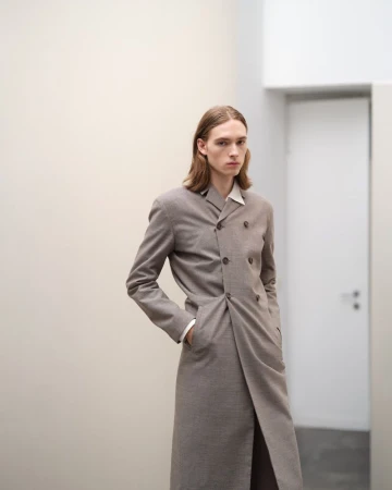 Model wearing a long taupe double-breasted coat standing in a minimalist interior space for Jil Sander.