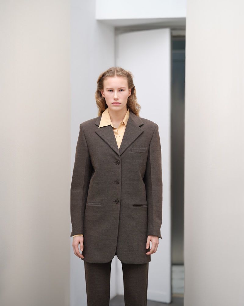 Woman in a brown suit and yellow shirt posing in a hallway with white walls and doorway for a fashion editorial style photograph.