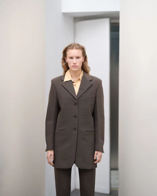 Woman in a brown suit and yellow shirt posing in a hallway with white walls and doorway for a fashion editorial style photograph.