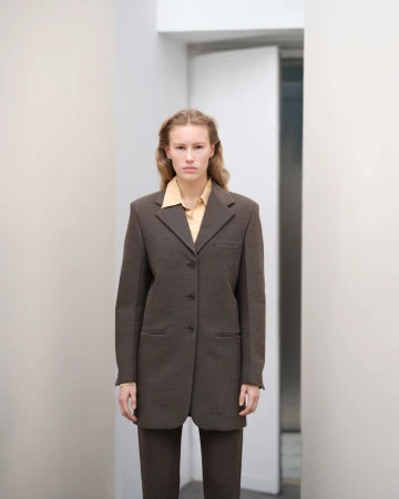 Woman in a brown suit and yellow shirt posing in a hallway with white walls and doorway for a fashion editorial style photograph.