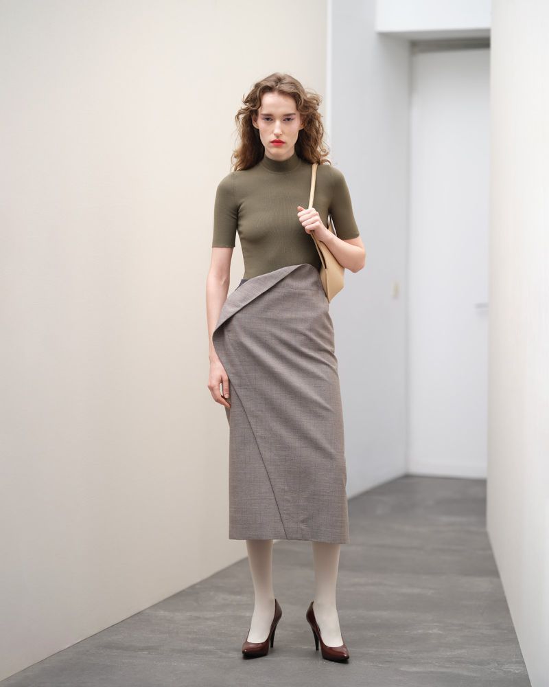 Adult female fashion model in a studio wearing an olive top, high-waisted skirt with a tan handbag and brown high heels.