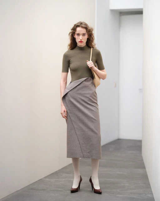 Adult female fashion model in a studio wearing an olive top, high-waisted skirt with a tan handbag and brown high heels.
