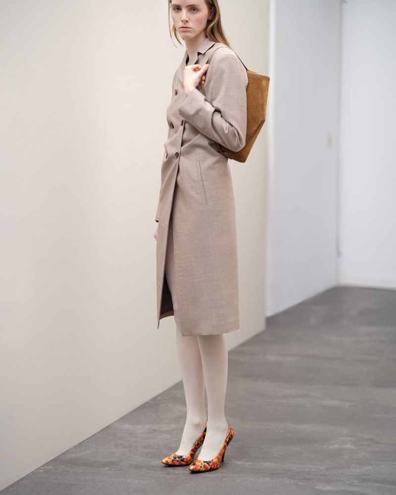 Fashion shot of a young adult female wearing a beige coat dress, white tights, and floral print heels, carrying a brown suede bag.