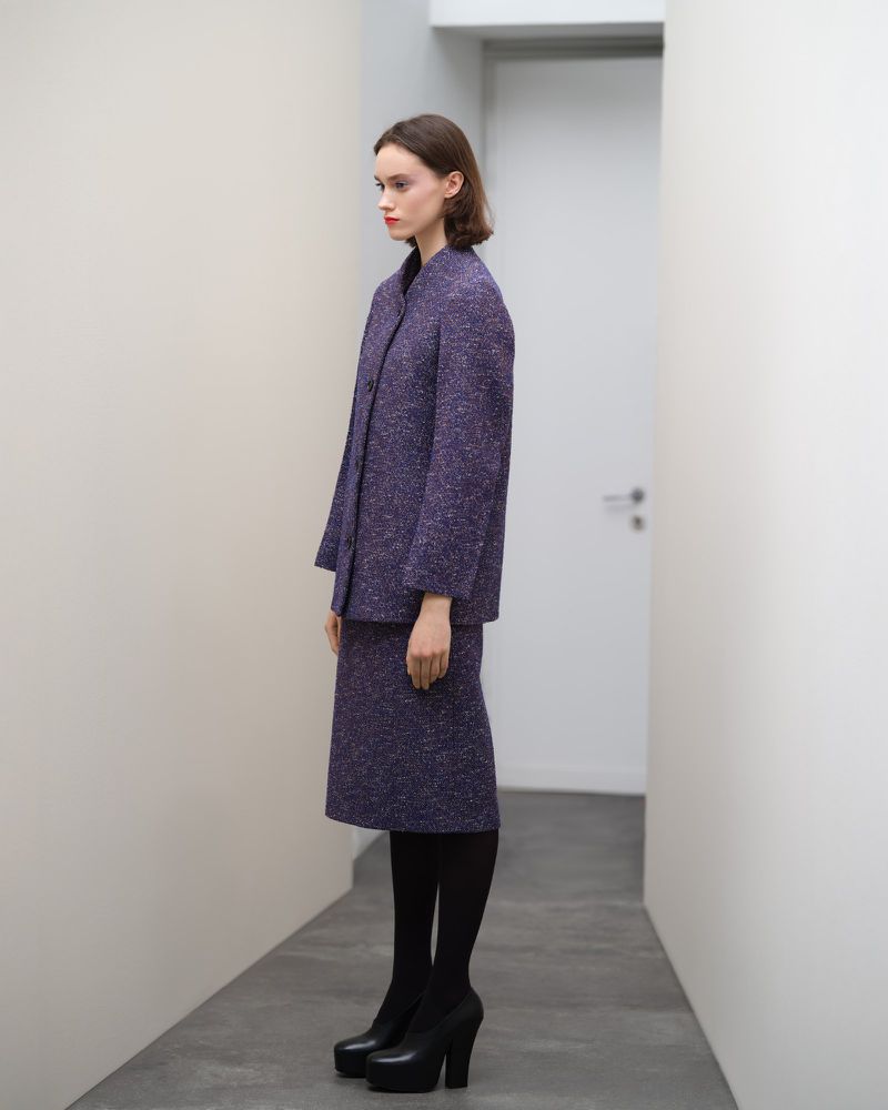 Adult female in a purple tweed suit and black platform heels stands in a bright, white hallway, side profile.