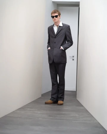 Model wearing dark sunglasses, grey suit jacket, white shirt, and trousers in a narrow hallway