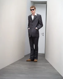 Model wearing dark sunglasses, grey suit jacket, white shirt, and trousers in a narrow hallway