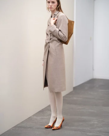 Model wearing a long beige belted coat and patterned heels holding a brown bag against a plain wall.