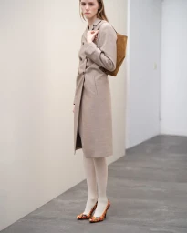 Model wearing a long beige belted coat and patterned heels holding a brown bag against a plain wall.