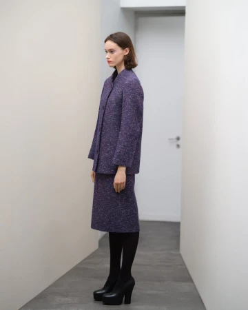 Model standing in profile wearing a purple tweed jacket and skirt set in a narrow white corridor