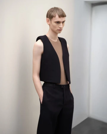 Model wearing a structured black vest over a brown top and black trousers against a plain wall.