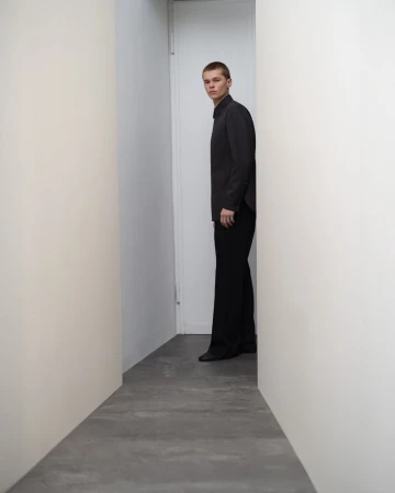 A young man wearing a black shirt and trousers standing in a narrow corridor
