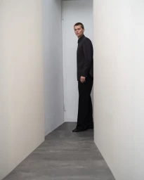 A young man wearing a black shirt and trousers standing in a narrow corridor