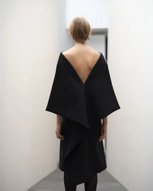 Model seen from behind wearing a structured black dress with a deep V-back in a minimalist white interior hallway