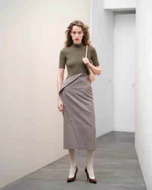 Model wearing an olive green short sleeve top and taupe midi skirt standing in a minimal interior hallway