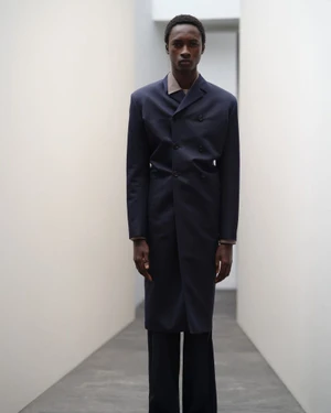 A Black man standing in a narrow minimalist corridor wearing a navy double-breasted coat and a beige collar shirt.