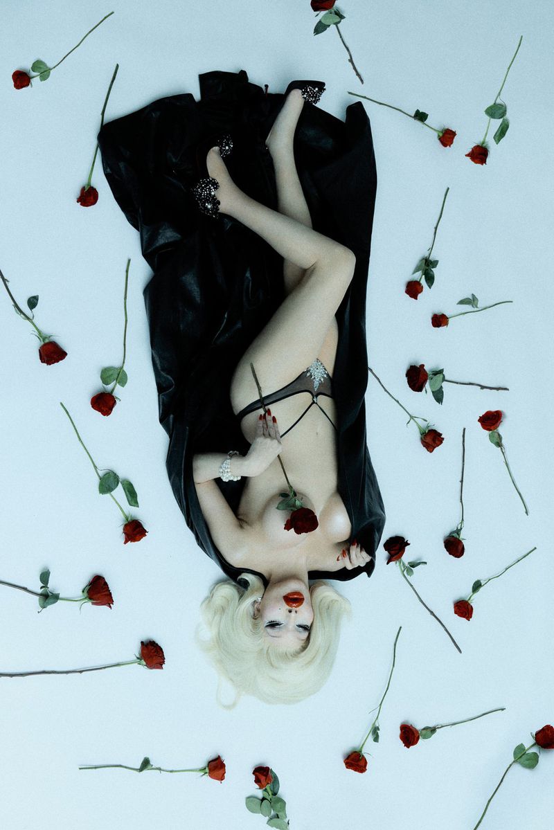 Overhead shot of a drag queen in lingerie lying on a white surface with red roses scattered around and wearing black high heels.