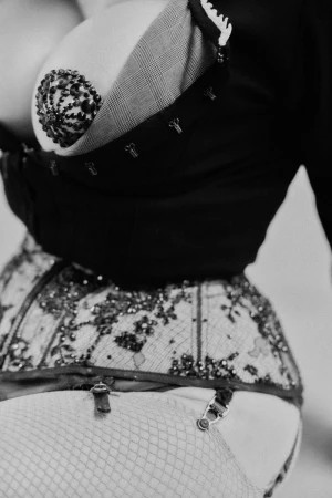 Close-up of a woman wearing a black corset with floral embellishments and fishnet stockings, posing in a studio with monochromatic tones.