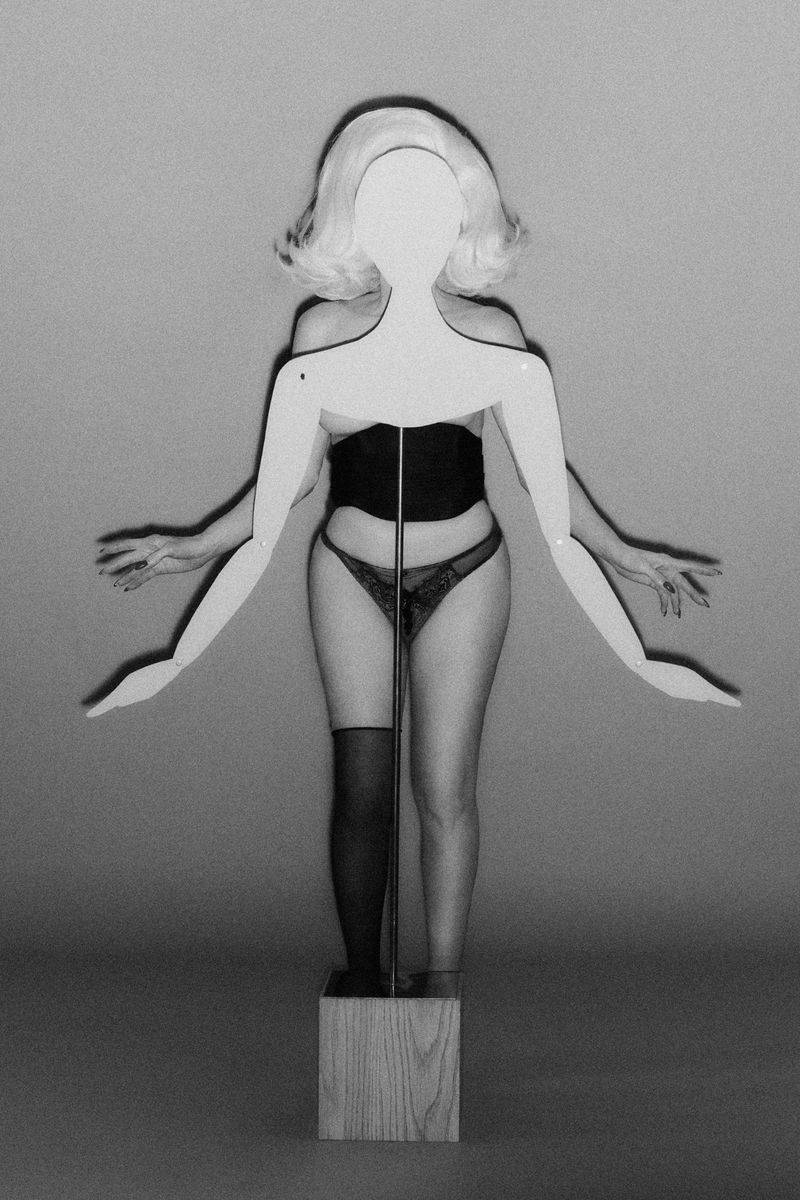 Abstract studio shot of a woman in lingerie standing behind a mannequin with arms outstretched, creating a multi-armed effect against a gray backdrop.