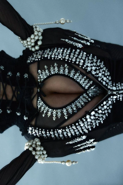 Close-up of a female torso wearing an ornate black top featuring embellishments, sheer fabric, and pearl jewelry against a light background.
