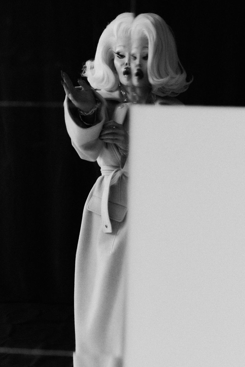 Black and white portrait of a person in a pale coat with a belt, wearing a large blonde wig and dramatic makeup against a dark background.