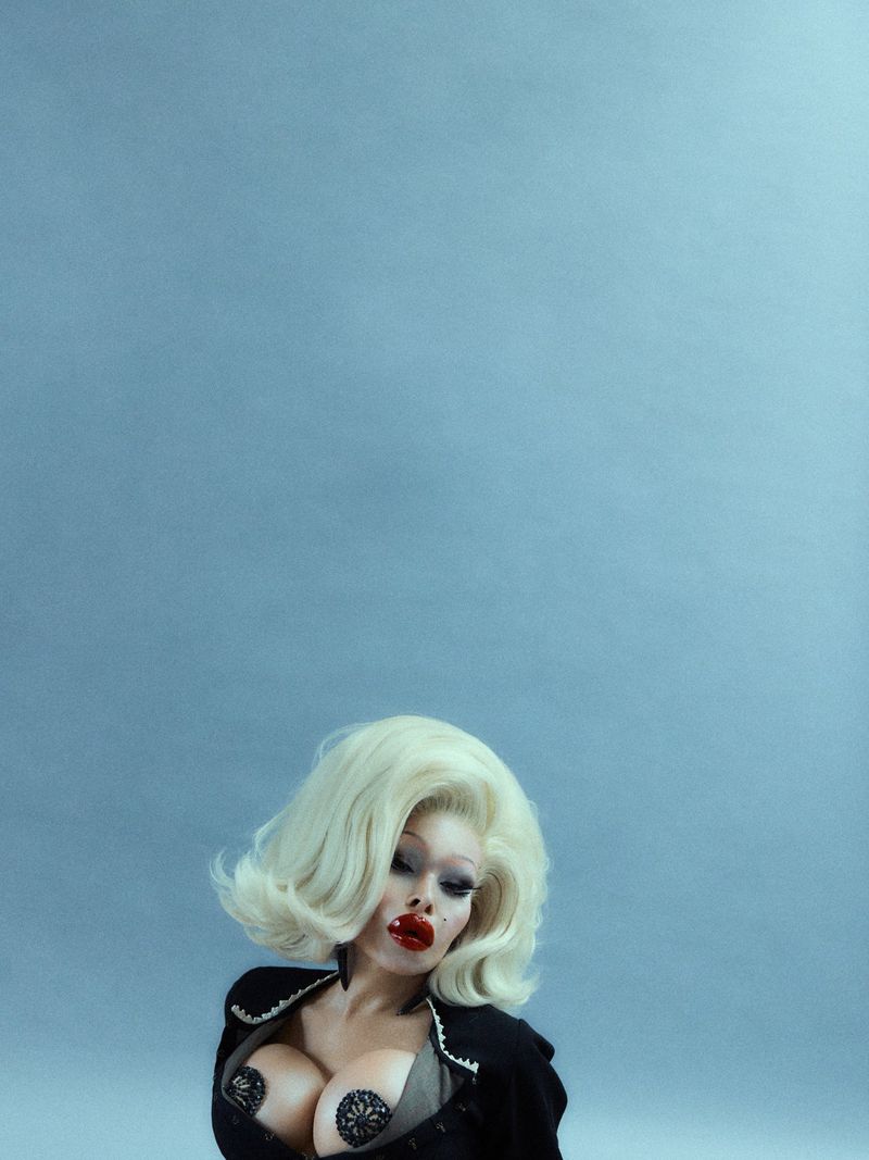 Adult female with white blonde hair poses wearing a black jacket against a solid blue background in a studio environment.