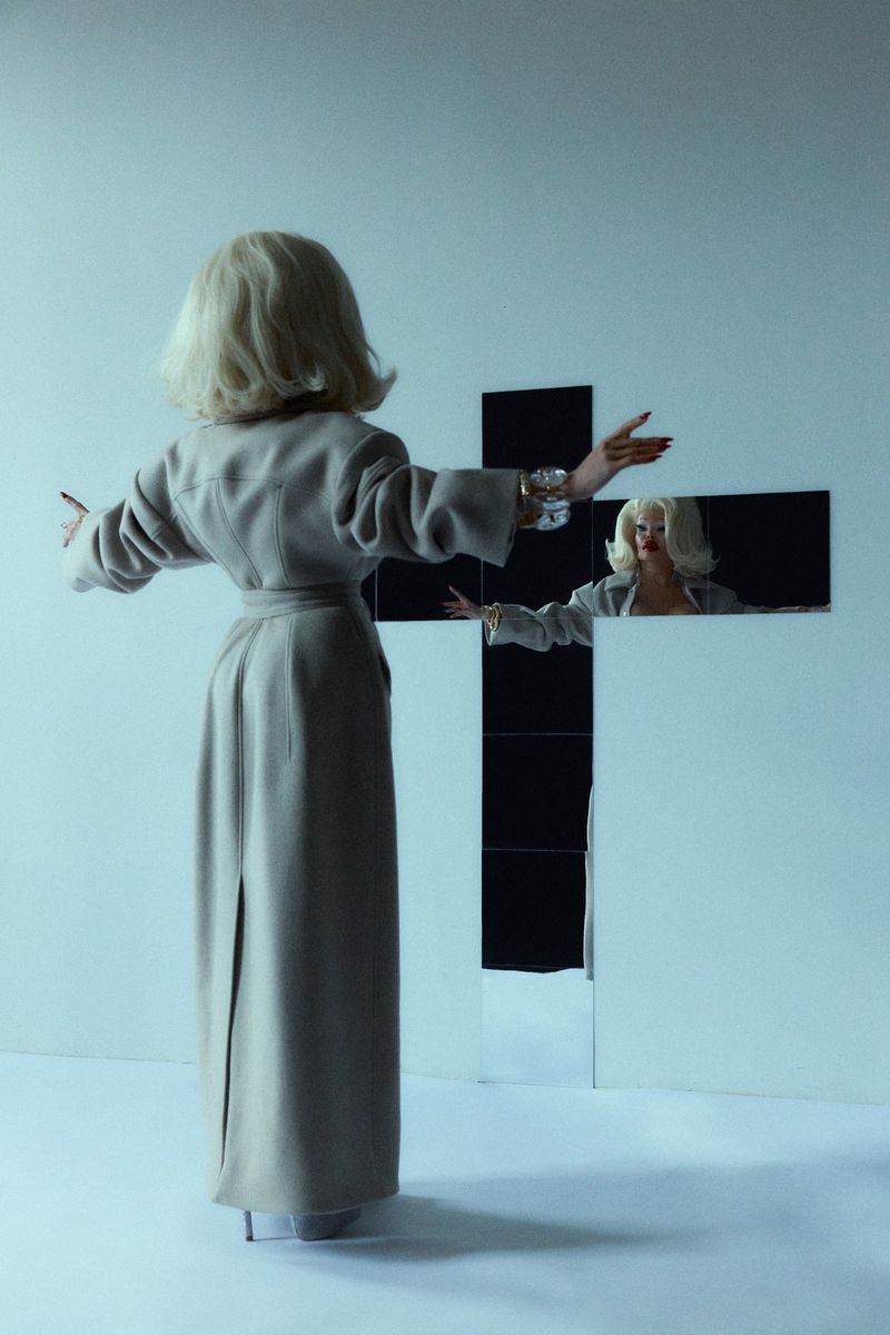 Blonde woman in a beige coat poses in a light blue room with her back to the viewer, in front of a cross constructed of mirrors.