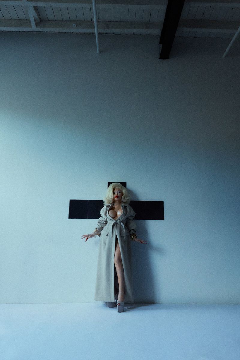Blonde woman in a trench coat stands in front of a cross in a photo studio with concrete floors and white walls, striking a pose.