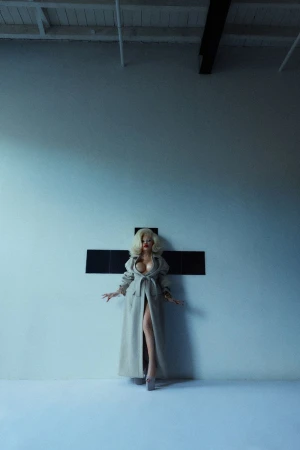 Blonde woman in a trench coat stands in front of a cross in a photo studio with concrete floors and white walls, striking a pose.