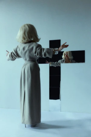Amanda Lepore viewed from behind wearing a beige belted coat facing a cross-shaped mirror against a white seamless background