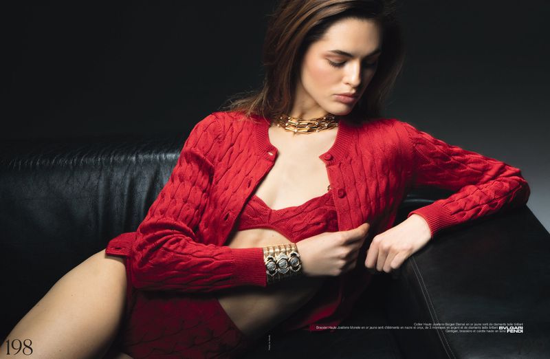 A woman in a red bra and cardigan sits on a black leather couch against a dark background, wearing gold jewelry and a bracelet.