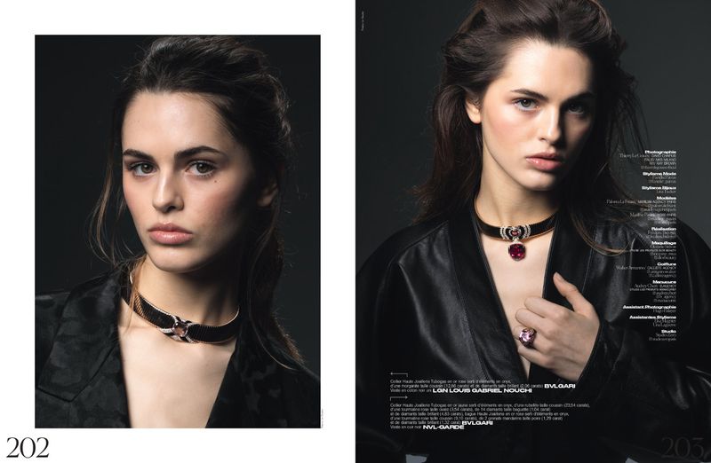 A young woman models a black choker necklace with a red ruby, wearing a black jacket, in a close-up studio shot.
