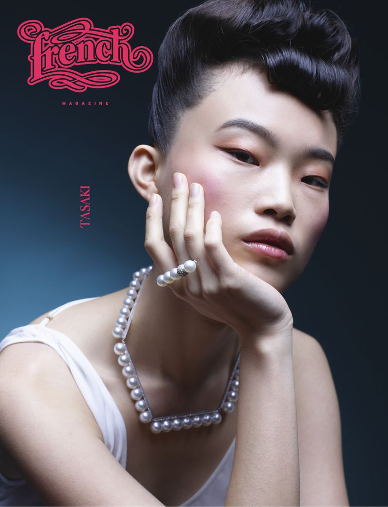 Asian woman with retro hairstyle poses for a fashion magazine cover, wearing pearl jewelry and a white top against a dark backdrop.
