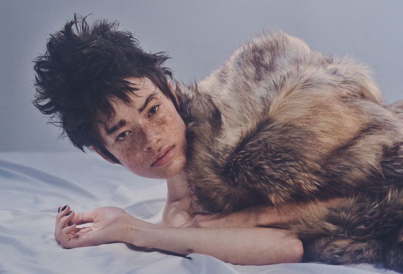 Portrait of a young adult person with freckles, dark hair, and a fur stole lying on a white sheet against a neutral background.