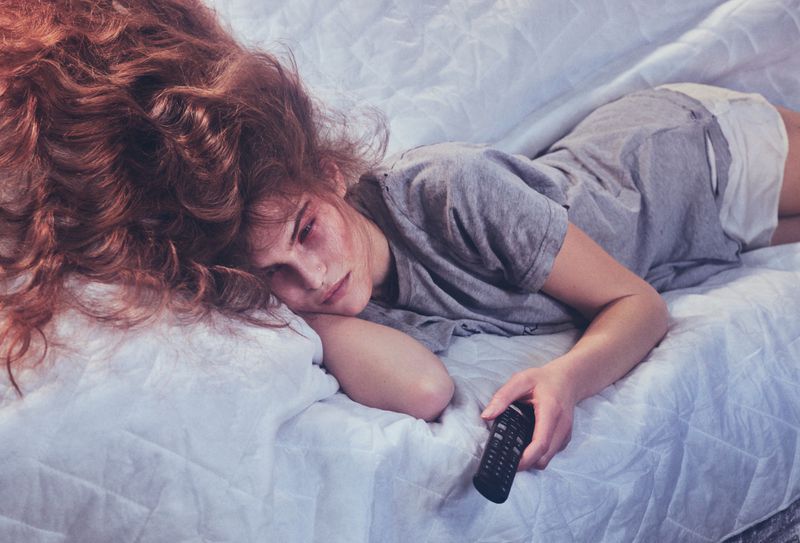 Woman lying on a white quilted bed with long red hair, holding a black television remote control, wearing a gray shirt and white shorts.