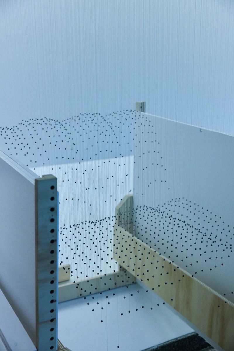 Abstract art installation featuring a box-like structure with strings and black dots, set against a white wall in an indoor setting.