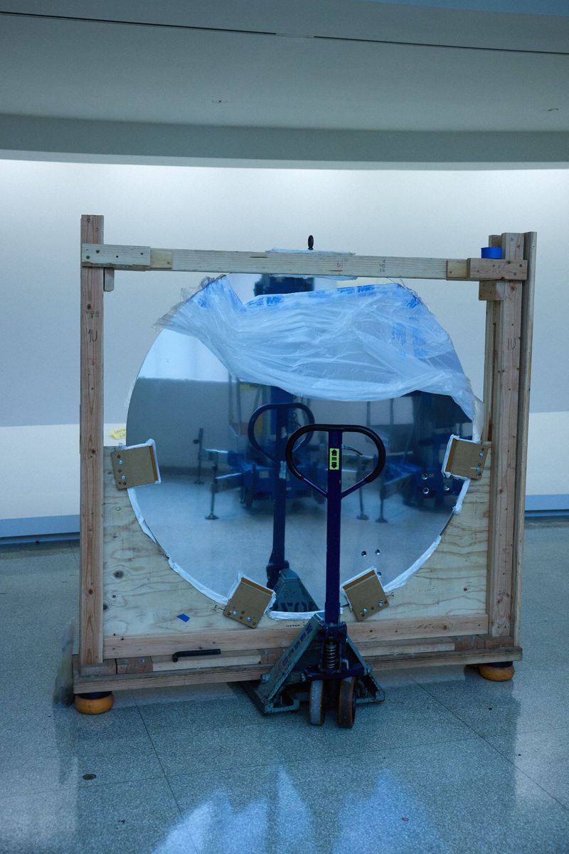 Large round mirror secured in a wood crate and placed on a blue dolly in a bright indoor space with gray tile flooring.