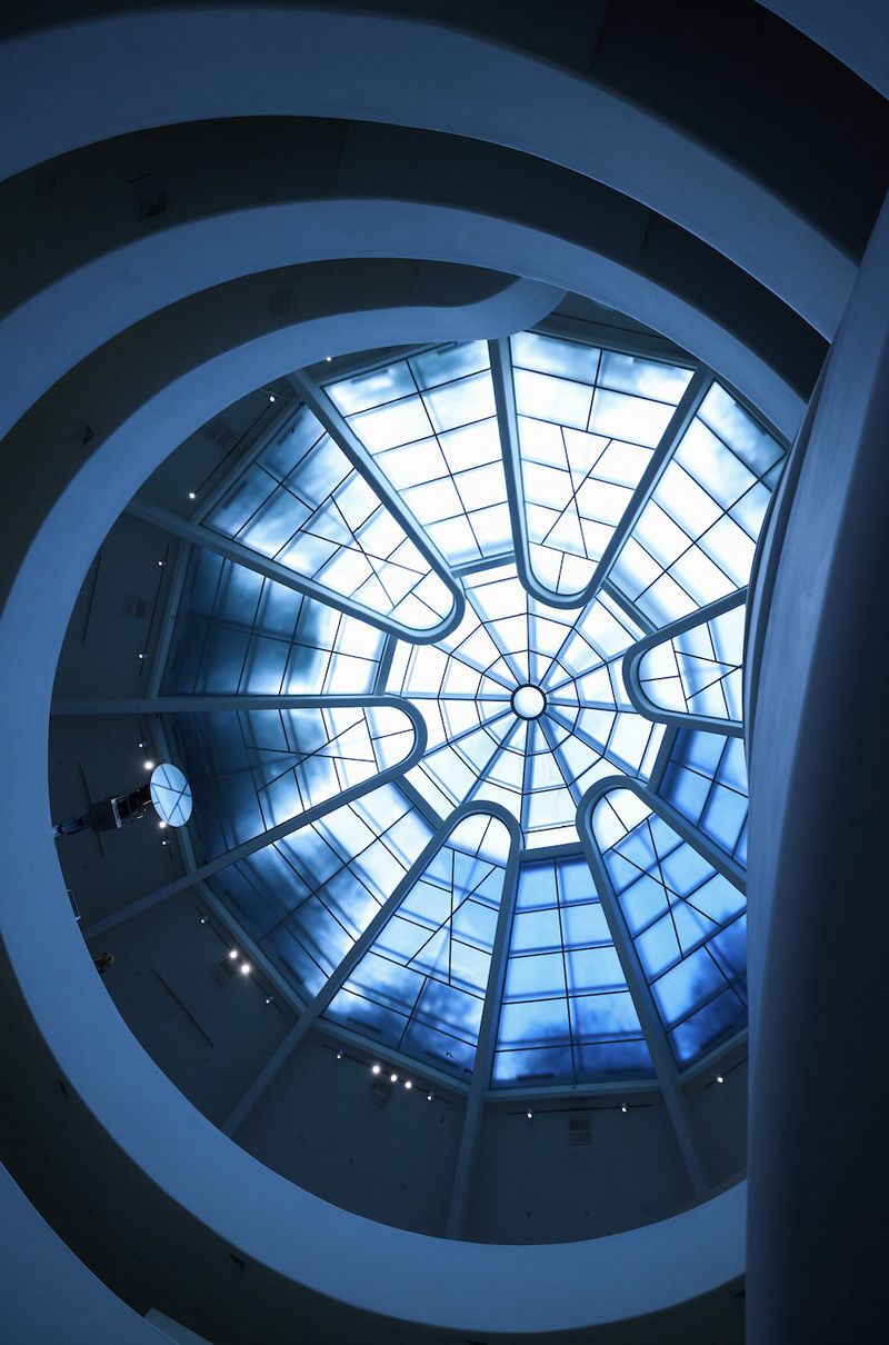 Looking up at a circular glass ceiling with geometric patterns and concrete architecture in an indoor setting with blue tones.