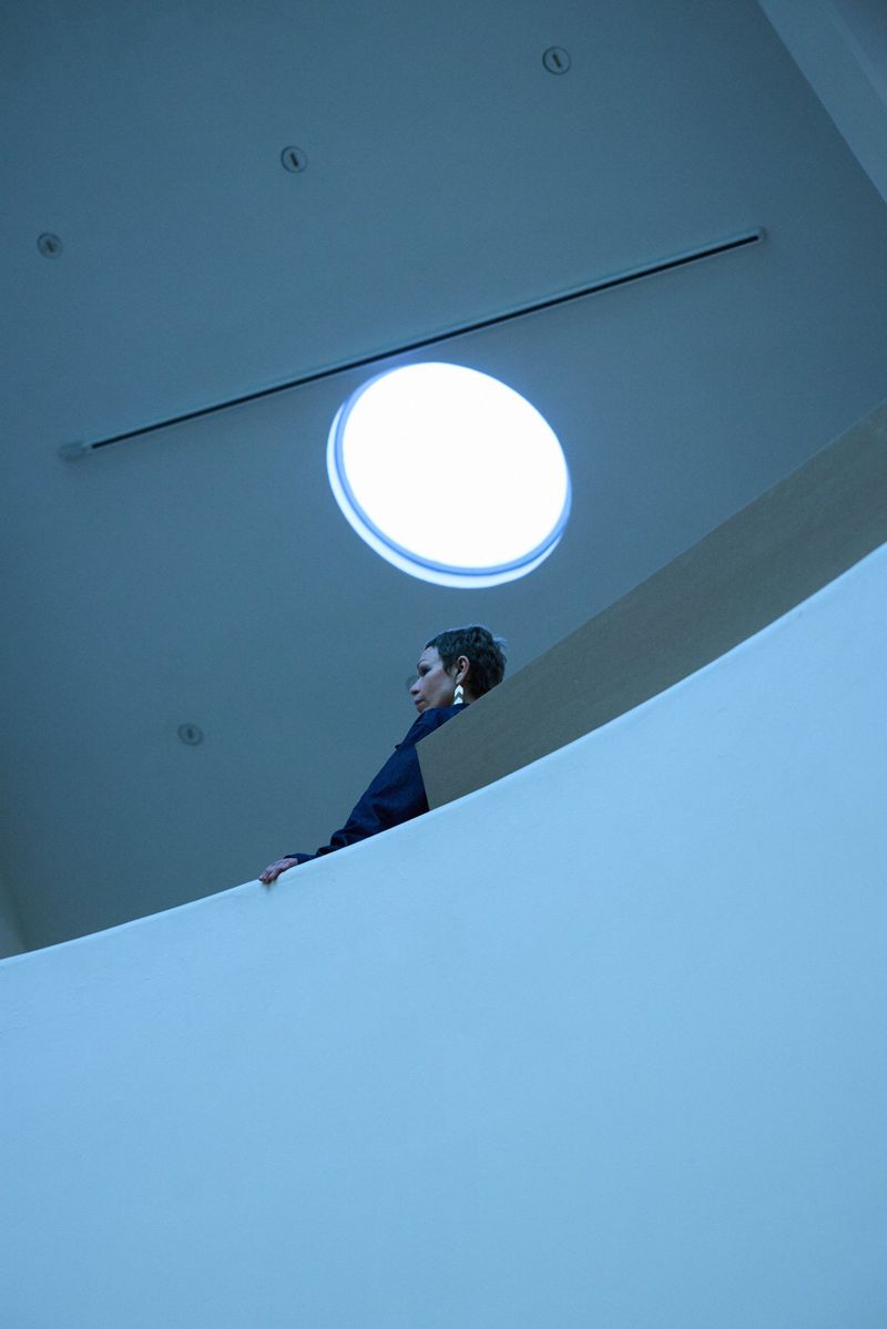 Woman in a navy jacket poses near a curved white wall under a round skylight in a minimalist indoor setting with blue lighting.
