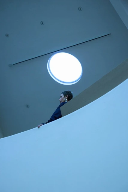 Woman in a navy jacket poses near a curved white wall under a round skylight in a minimalist indoor setting with blue lighting.