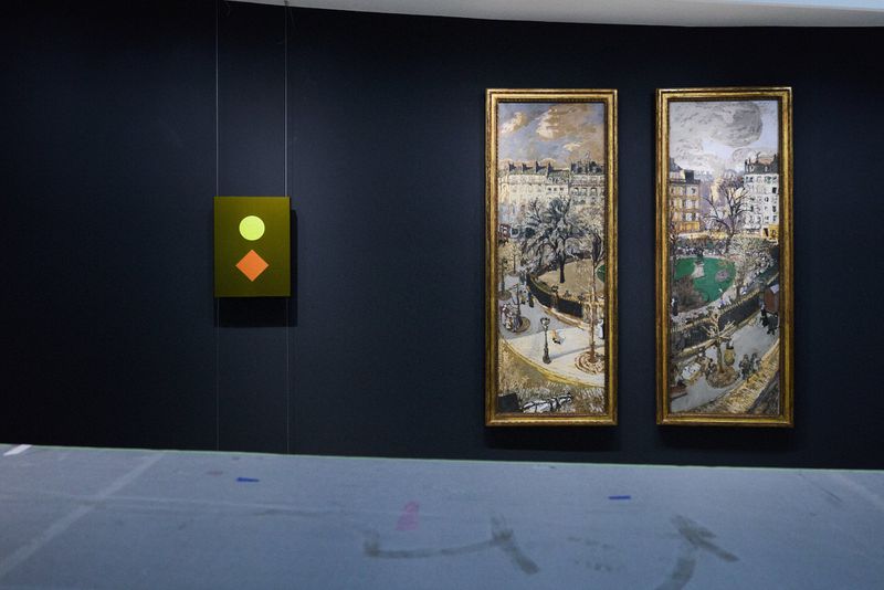 Indoor shot of framed art pieces on a dark wall, including cityscapes and abstract geometric art, displayed in an art gallery.